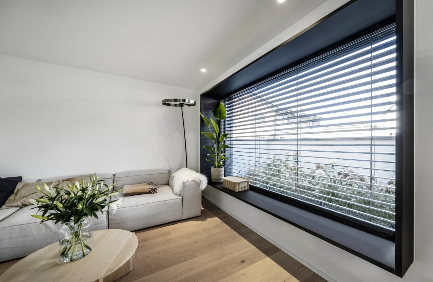 Modernes Wohnzimmer mit grauem Sofa, Pflanzen und großen Fenstern mit Jalousien